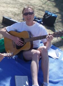 Doctor Rubato is sitting on a blanket on the lawn near the Chautauqua auditorium in Boulder. He is playing an acoustic guitar, which rests on his right leg, and appears to be singing. He is wearing sunglasses, a t-shirt, shorts, and socks, although his feet are out of frame. Papers and a book are on the blanket near him, presumably with music. Behind him on the lawn is a guitar case, a carrying bag, and some other identifiable items, possibly a jacket or other clothing or a backpack. The scene is mostly sunny, although there is some shade from a tree which is not in the picture. To his right is the end of a guitar and a hand, presumably belonging to another guitarist.