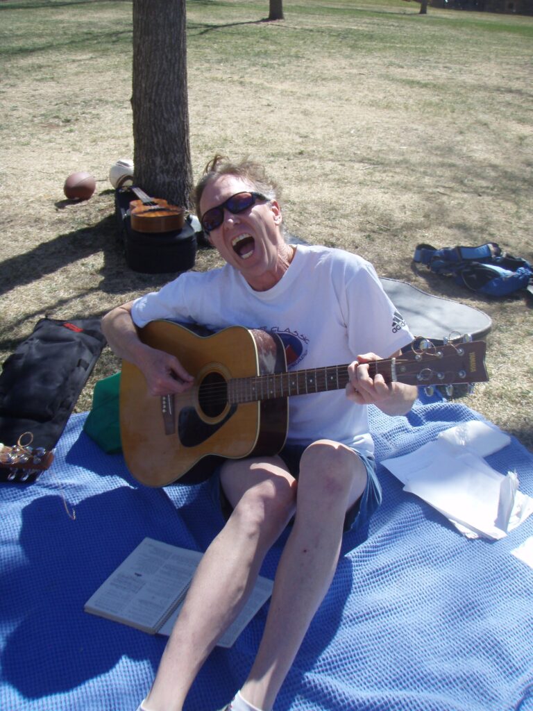 Doctor Rubato is sitting on a blanket on the lawn near the Chautauqua auditorium in Boulder. He is playing an acoustic guitar, which rests on his right leg, and appears to be singing enthusiastically. He is wearing sunglasses, a t-shirt, shorts, and socks, although his feet are out of frame. Papers and a book are on the blanket near him, presumably with music. Behind him on the lawn to his left is a guitar case and a small backpack. Behind him is a the trunk of a tree. Under the tree is another musical instrument, possibly a ukulele, resting on its case. Also under the tree are a couple of balls, possibly a football and a soccer ball. Next to him on his right is a soft case likely for a small guitar. The scene is mostly sunny, although there is some shade from one or more unseen trees. To his right is the end of a guitar, presumably belonging to another guitarist.